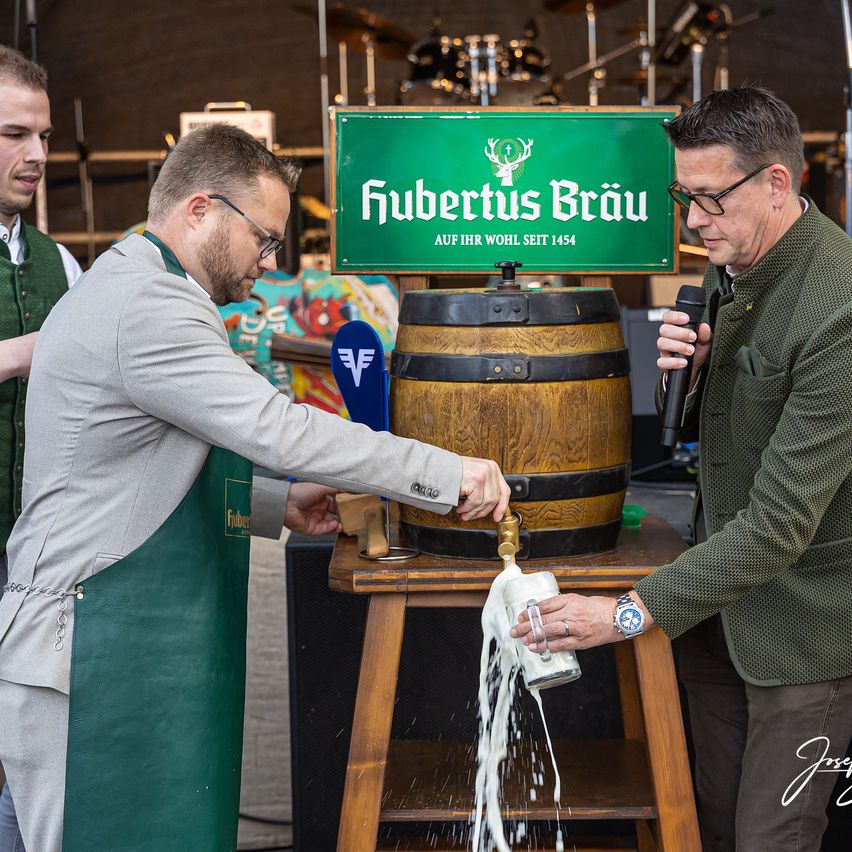 Zwei Männer schenken Bier aus einem Holzfass mit der Aufschrift 'Fubertus Brau' ein. Ein Mann hält ein Glas, während der andere den Zapfhahn hält. Ein dritter Mann steht hinter ihnen mit einem Mikrofon.