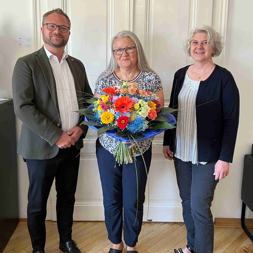 Drei Personen, zwei Frauen und ein Mann, stehen lächelnd zusammen in einem Raum. Die Frau in der Mitte hält einen großen Strauß bunter Blumen.