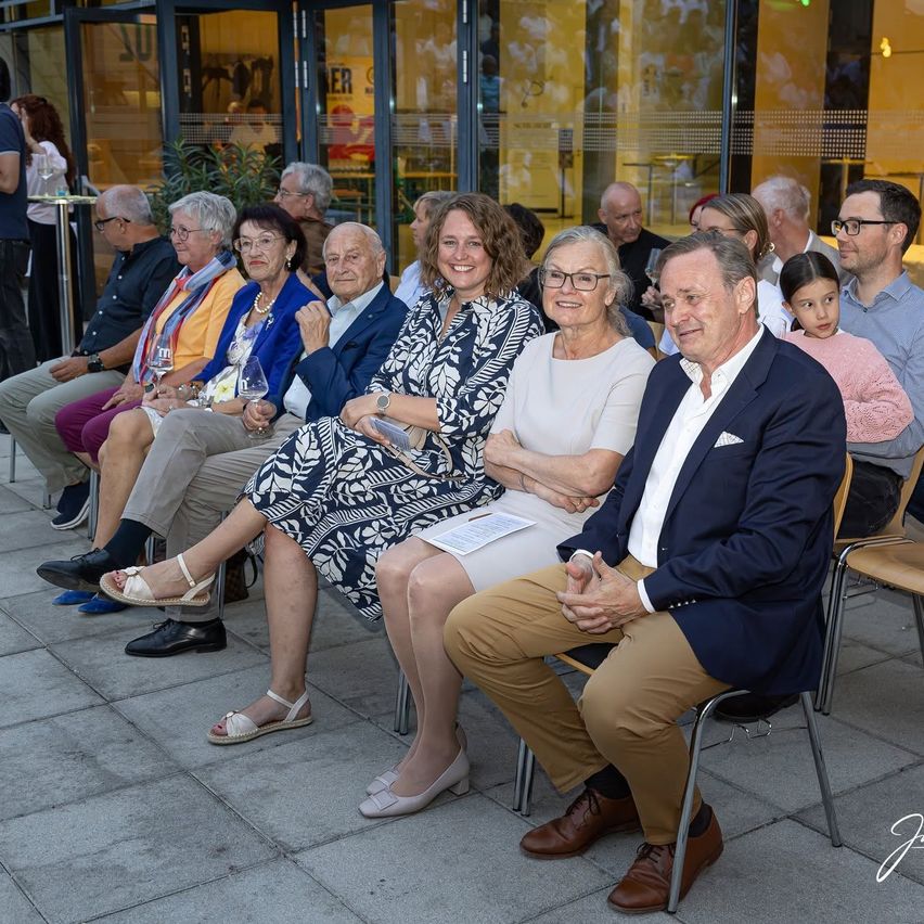 Eine Gruppe älterer Menschen sitzt auf Stühlen vor einem Gebäude und lächelt. Sie scheinen an einer sozialen Veranstaltung teilzunehmen.
