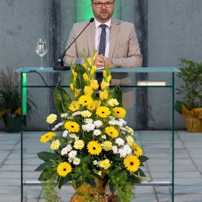 Ein Mann in einem Anzug steht hinter einem Podium mit einem Mikrofon, umgeben von einem großen Strauß gelber und weißer Blumen.