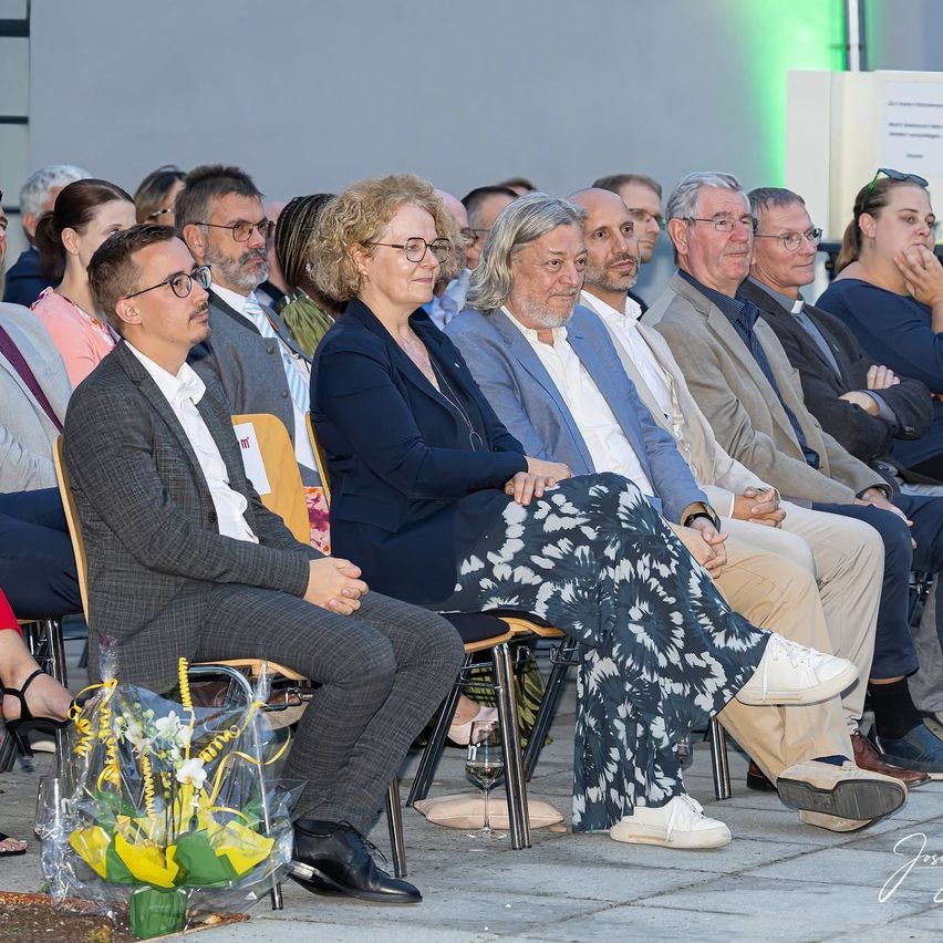 Eine Gruppe von Menschen sitzt auf Stühlen, wahrscheinlich bei einer formellen Veranstaltung. Einige tragen Brillen und Anzüge. Eine Frau in der Mitte hat ein Blumenarrangement vor sich.