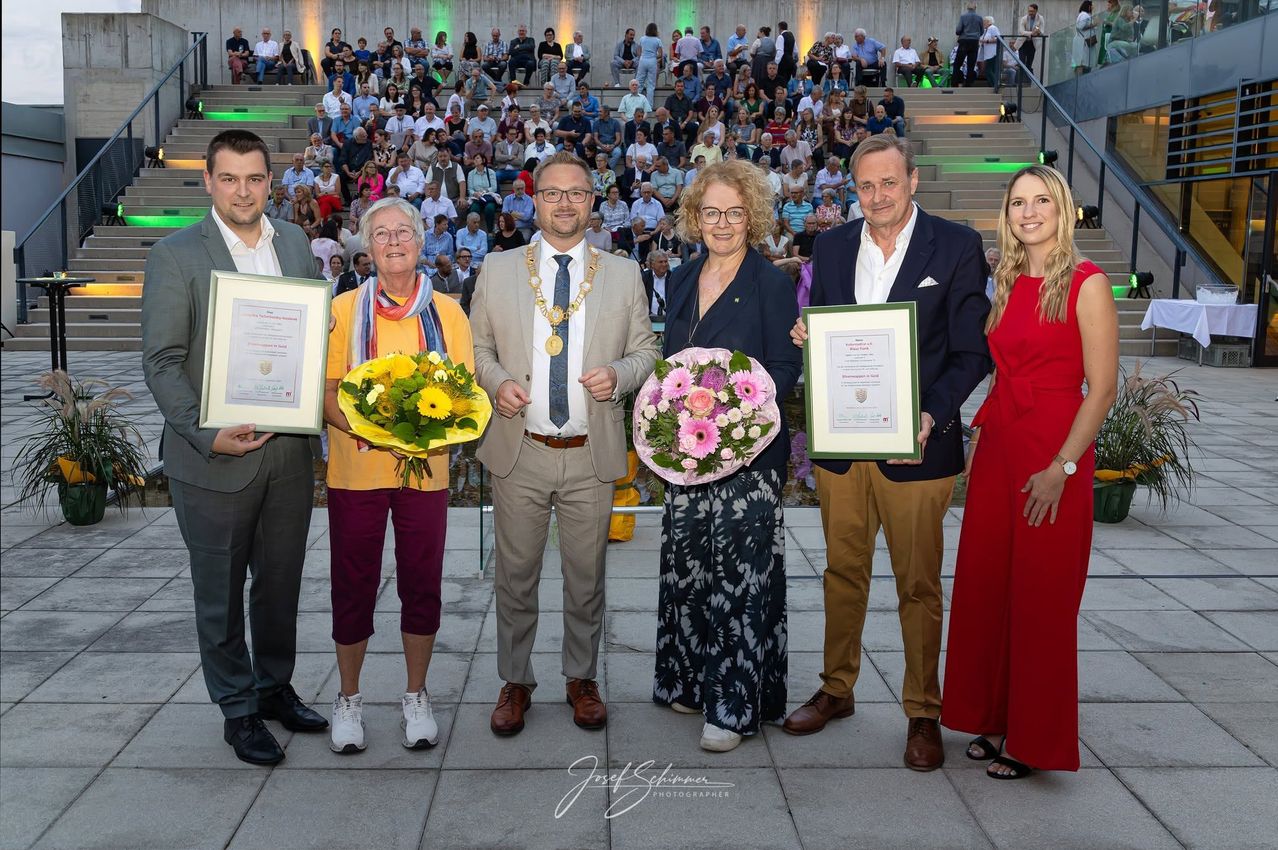 Eine Gruppe von Menschen, die zusammenstehen, Urkunden und Blumen vor einem Publikum halten.