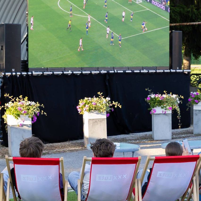 Drei Kinder sitzen in roten und weißen Stühlen und schauen ein Fußballspiel auf einer großen Leinwand mit Motorola-Logo. Die Leinwand zeigt Spieler in blauen und weißen Trikots, mit Lautsprechern und Topfpflanzen im Hintergrund.