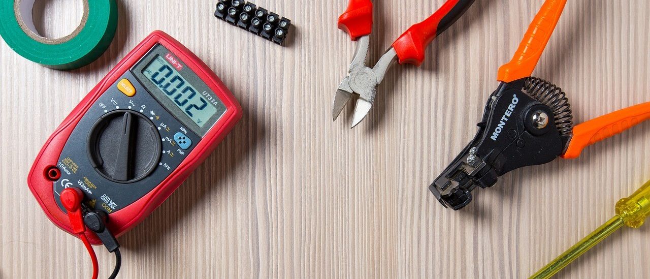 Auf einem Holztisch sind verschiedene Werkzeuge zu sehen: ein digitales Multimeter, ein roter und schwarzer Schraubendreher, ein Paar Drahtschneider, ein Paar Zangen, eine Rolle Klebeband und einige Kabel.