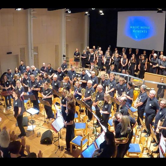 Ein großes Ensemble von Musikern, einschließlich Blechbläser, Holzbläser und Percussion, spielt auf der Bühne. Im Hintergrund zeigt eine große Leinwand 'Magic Movie Moments'.