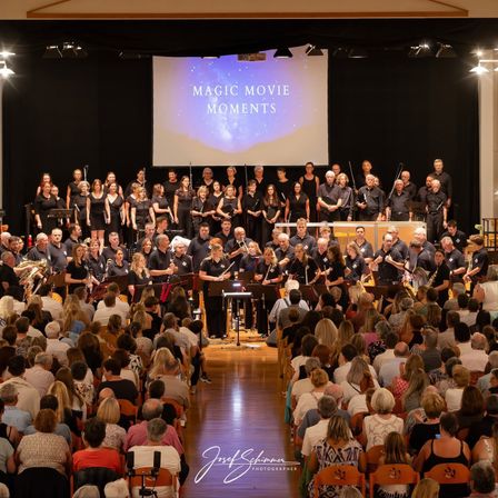 Ein großes Ensemble von Musikern tritt auf der Bühne vor einem großen Publikum auf, wobei eine Projektionsleinwand hinter ihnen 'Magic Movie Moments' anzeigt.