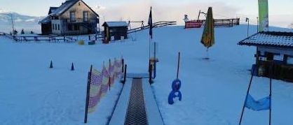 Verschneite Skipiste mit einem Skilift, einem Haus und einem kleinen Gebäude. Fahnen und Banner sind auf der Piste platziert.