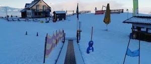 Verschneite Skipiste mit einem Skilift, einem Haus und einem kleinen Gebäude. Fahnen und Banner sind auf der Piste platziert.