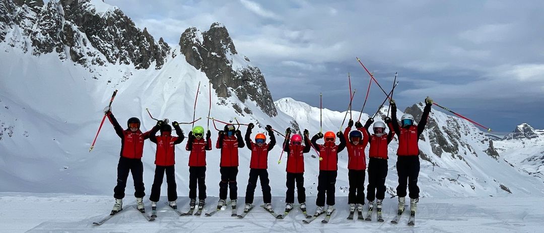 Eine Gruppe von Skifahrern in roten Uniformen posiert für ein Foto auf einem verschneiten Berg. Sie halten Skistöcke und tragen Helme.