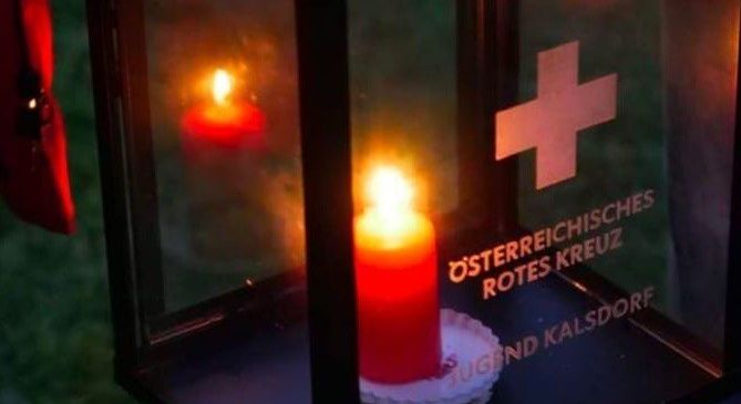 A lantern with a lit candle inside and a red cross symbol. The text 'Osterreichisches Rotes Kreuz' and 'Friedenslicht' is on the lantern.