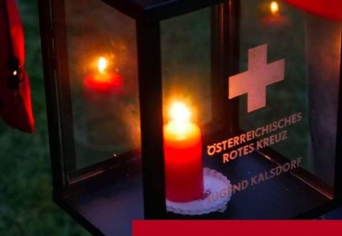Eine Laterne mit einer brennenden Kerze im Inneren und einem roten Kreuz-Symbol. Der Text 'Osterreichisches Rotes Kreuz' und 'Friedenslicht' steht auf der Laterne.