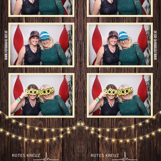 Eine Fotobox-Collage zeigt vier Bilder von zwei Frauen in grünen Kleidern, eine trägt einen blauen Hut und eine Brille, die andere einen braunen Hut. Der Hintergrund hat rote Laternen und eine Holzwand.