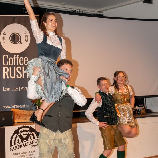 Eine Frau wird von zwei Männern in traditioneller Kleidung bei einer Coffee Rush-Veranstaltung gehoben, mit einer Projektionsleinwand im Hintergrund.