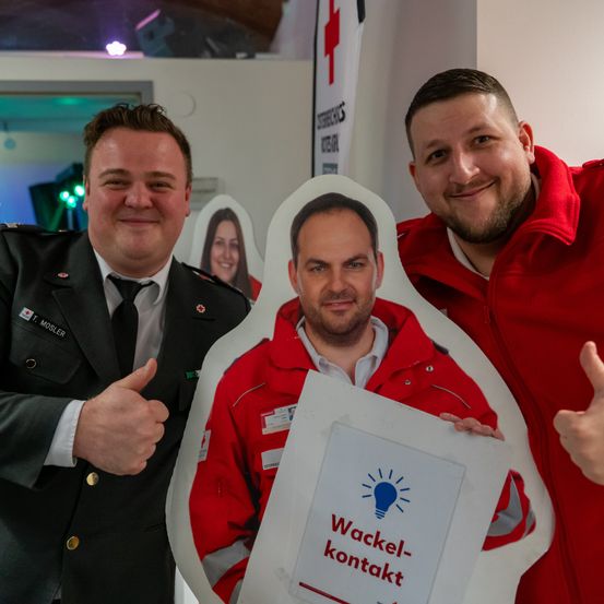 Zwei Männer in Uniformen und ein lächelnder Mann in einem roten Jackett posieren mit einer Ausschnitt eines Mannes, der ein Schild mit der Aufschrift 'Wackel-kontakt' hält.