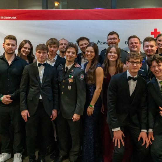 Eine Gruppe von Menschen in formeller Kleidung und Militäruniformen steht zusammen vor einem Banner. Sie scheinen für ein Foto zu posieren.