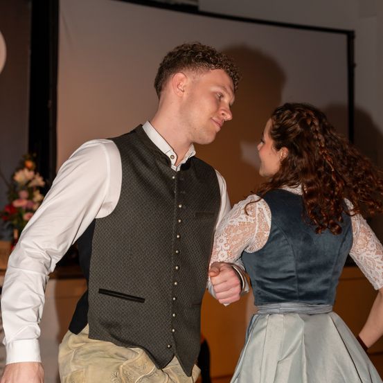 Ein Mann und eine Frau in traditioneller Kleidung tanzen zusammen, einander gegenüberstehend und halten Händchen. Die Frau hat geflochtene Haare. Hinter ihnen befindet sich eine weiße Wand mit einem Bildschirm.