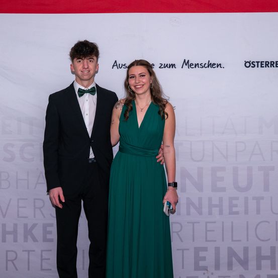 Ein Mann und eine Frau stehen bei einer Veranstaltung eng beieinander und lächeln. Der Mann trägt einen schwarzen Anzug und eine Fliege. Die Frau trägt ein grünes Kleid und hat eine Tätowierung am Arm. Hinter ihnen befindet sich ein weißer Hintergrund mit Text.