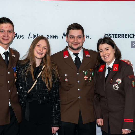 Vier Personen, zwei Männer und zwei Frauen, stehen lächelnd zusammen in Uniformen. Sie scheinen sich bei einer Veranstaltung zu befinden, mit einem weißen Hintergrund hinter ihnen.