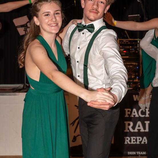 Ein junges Paar tanzt eng auf einer Bühne, lächelt und hält sich an den Händen. Die Frau trägt ein grünes Kleid und der Mann ein weißes Hemd mit grünen Hosenträger und eine Fliege.