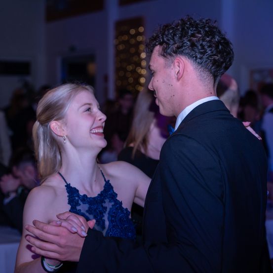 Ein Paar tanzt fröhlich auf einer Party. Die Frau lächelt hell, während der Mann sie mit einem zufriedenen Ausdruck ansieht. Der Hintergrund ist verschwommen, mit Leuten an Tischen und einer festlichen Atmosphäre.