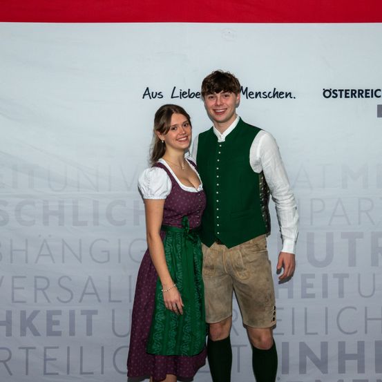 Ein lächelndes Paar in traditioneller Kleidung steht vor einem Banner mit deutschem Text. Der Mann trägt eine grüne Weste und braune Hose, und die Frau trägt ein lila Kleid. Sie sind wahrscheinlich bei einer Veranstaltung.