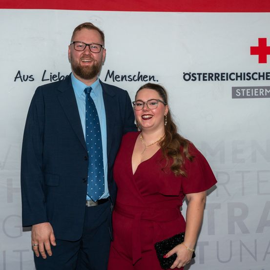 Ein Mann und eine Frau stehen nah beieinander, lächelnd, vor einer weißen Wand mit Text und rotem Kreuz. Sie tragen formelle Kleidung.