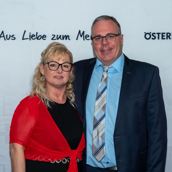Ein Mann und eine Frau stehen lächelnd eng beieinander vor einem weißen Hintergrund mit schwarzem Text, der 'Aus Liebe zum Me' und 'oSTER' lautet. Die Frau trägt einen roten Schal und der Mann ein blaues Hemd und eine Krawatte.