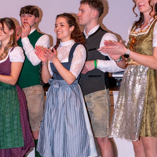 Eine Gruppe von Menschen, wahrscheinlich Schauspieler, in traditioneller Kleidung klatscht und lächelt. Die Frau in der Mitte trägt ein blaues Kleid. Hinter ihnen dient eine weiße Wand als Hintergrund.