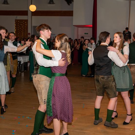 Mehrere junge Leute tanzen in traditioneller Kleidung bei einer Veranstaltung. Die Paare stehen eng beieinander, und der Raum ist mit Lichtern und Konfetti dekoriert.