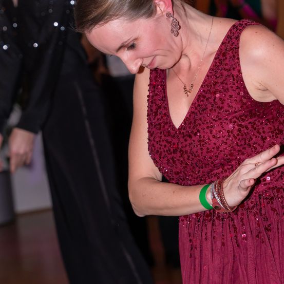 Eine Frau in einem Paillettenkleid in Weinrot tanzt und schaut nach unten. Hinter ihr steht eine weitere Person in Schwarz, und sie befinden sich in einem schwach beleuchteten Raum.
