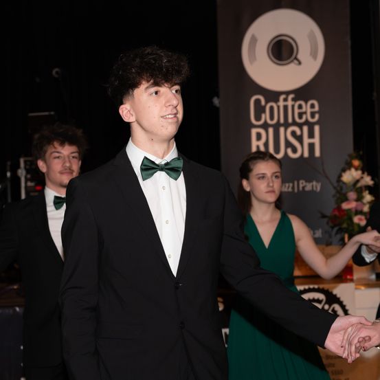 Ein junger Mann in einem schwarzen Frack und einer grünen Fliege steht mit einer Frau in einem grünen Kleid. Sie sind wahrscheinlich bei einer formellen Veranstaltung. Ein Banner hinter ihnen trägt den Text Coffee Rush.
