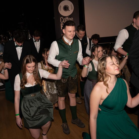 Eine Gruppe von Menschen tanzt auf einem Holzboden. Einige tragen traditionelle Kleidung. Ein Mann ist in einem grünen Vest, und zwei Frauen sind in grünen Kleidern.