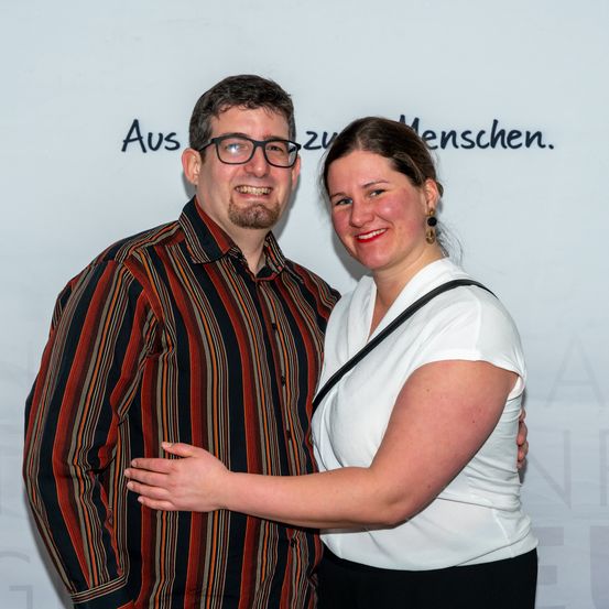 Ein Mann und eine Frau stehen eng beieinander mit umschlungenen Armen vor einer weißen Wand.
