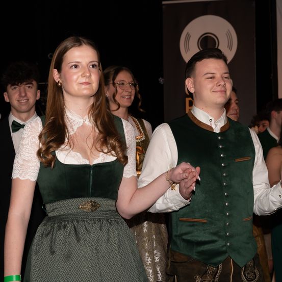Ein junges Paar in traditioneller deutscher Kleidung, lächelnd und Händchen haltend. Hinter ihnen sind mehrere Personen anwesend, einschließlich eines Mannes mit einer Fliege. Die Umgebung scheint ein festliches Ereignis zu sein.