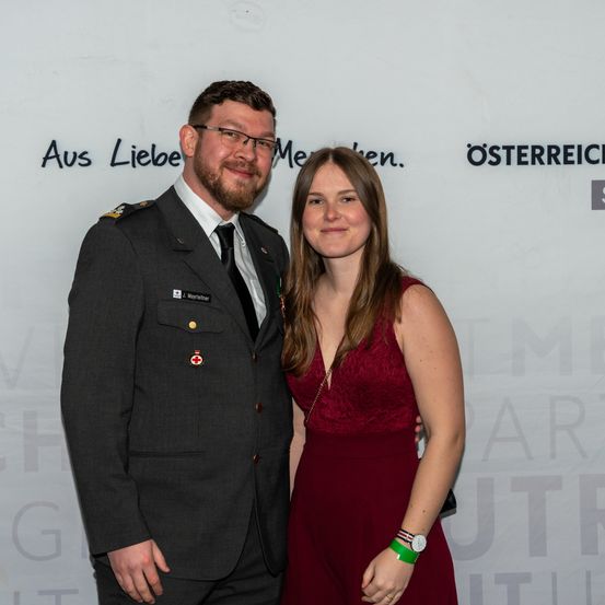 Ein Mann in Uniform und eine Frau in einem roten Kleid stehen zusammen und lächeln. Der Mann hat einen roten Kreuzabzeichen auf seiner Uniform. Hinter ihnen ist eine Wand mit deutschem Text.
