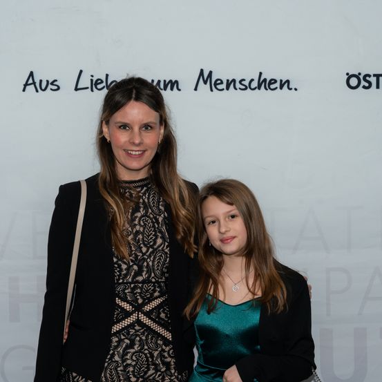 Zwei Frauen stehen nebeneinander, lächeln und posieren für ein Foto. Hinter ihnen ist eine weiße Wand mit schwarzem Text.