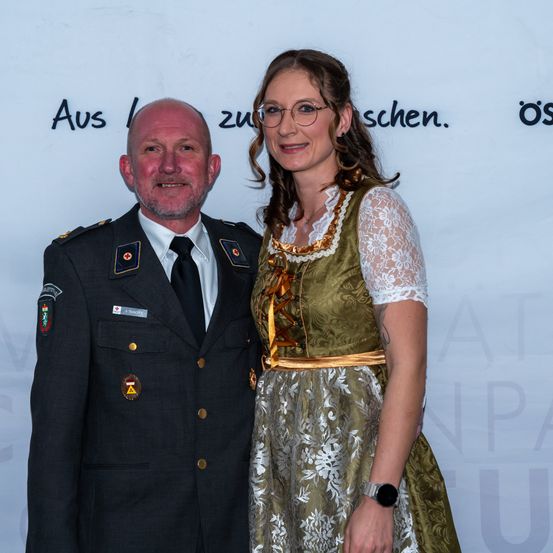 Ein Mann in Uniform und eine Frau in einem grünen Kleid stehen nebeneinander und lächeln für ein Foto. Dahinter zeigt eine weiße Hintergrundwand schwarzen Text.