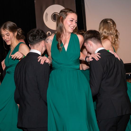 Eine Gruppe junger Menschen versammelt sich um eine Frau in einem grünen Kleid, lächelt und sieht glücklich aus. Die Frau trägt eine Halskette und hat ihre Arme um zwei junge Männer gelegt. Der Hintergrund ist unscharf, mit einem Banner und einer weißen Wand.