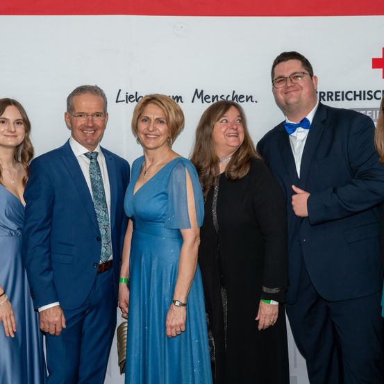 Eine Gruppe von formell gekleideten Menschen, darunter zwei Frauen in blauen und schwarzen Kleidern, posieren für ein Foto vor einem weißen Hintergrund mit der Aufschrift 'Liebe um Menschen.' Die Gruppe umfasst einen Mann mit einer blauen Schleife.