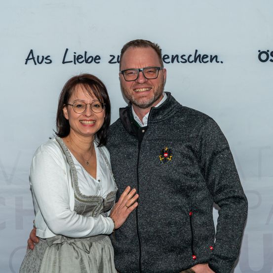 Ein lächelndes Paar steht vor einer weißen Wand mit schwarzem Text, die Frau trägt ein weißes Oberteil und eine Halskette, und der Mann trägt eine graue Jacke mit einem Logo auf der linken Seite.