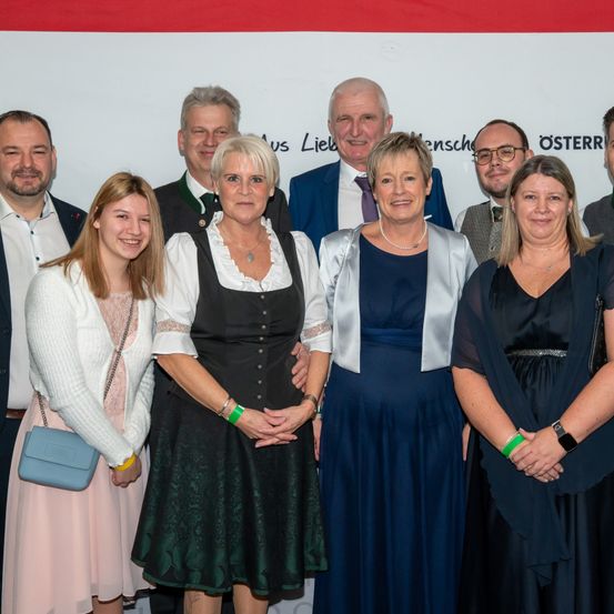 Eine Gruppe von Menschen, wahrscheinlich eine Familie, steht lächelnd für ein Foto zusammen. Sie stehen vor einem weißen Hintergrund mit Text. Die Menschen tragen verschiedene Kleidungsstücke.