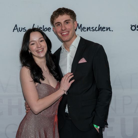 Ein junges Paar steht lächelnd vor einem weißen Hintergrund mit schwarzem Text 'Aus Liebe Menschen.' Die Frau trägt ein funkelnde Kleid und der Mann hat ein rosa Taschentuch in der Brusttasche seines Anzugs.