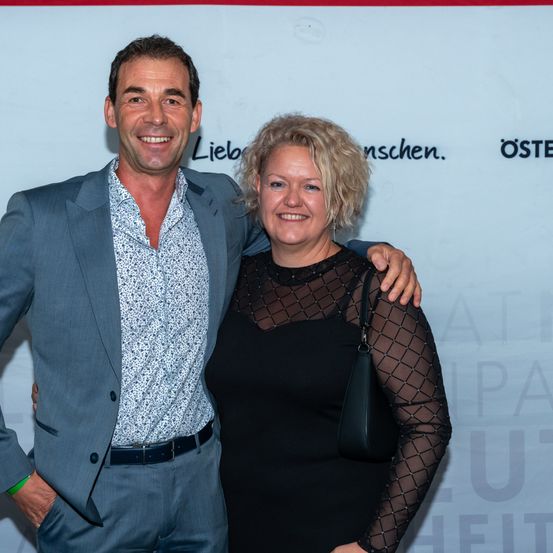 Ein Mann und eine Frau lächeln und posieren für ein Foto, stehen nah beieinander und haben die Arme umeinander geschlungen.