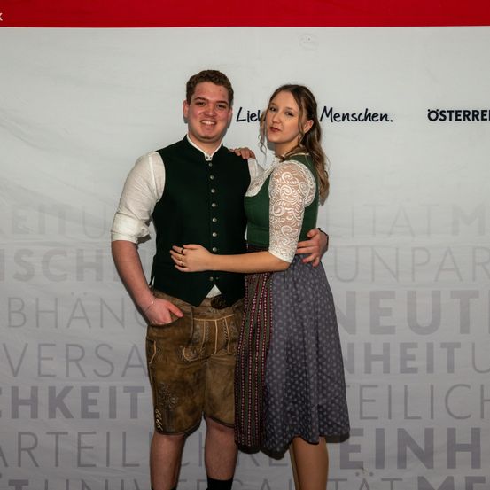 Ein Mann und eine Frau in traditioneller deutscher Kleidung stehen eng beieinander vor einem weißen Hintergrund mit Text.