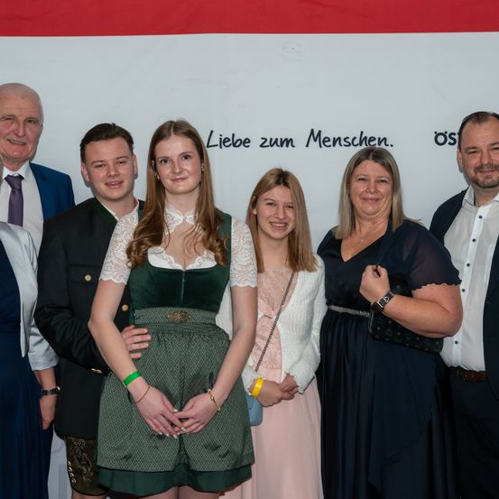 Eine Gruppe von Menschen in formeller Kleidung posiert für ein Foto vor einem weißen Hintergrund mit deutschem Text.