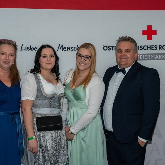 Vier Personen stehen vor einem Hintergrund mit Text und rotem Kreuz und lächeln für ein Foto. Die beiden Frauen tragen traditionelle Kleider, eine hält eine schwarze Clutch. Der Mann trägt einen Anzug und eine Fliege.