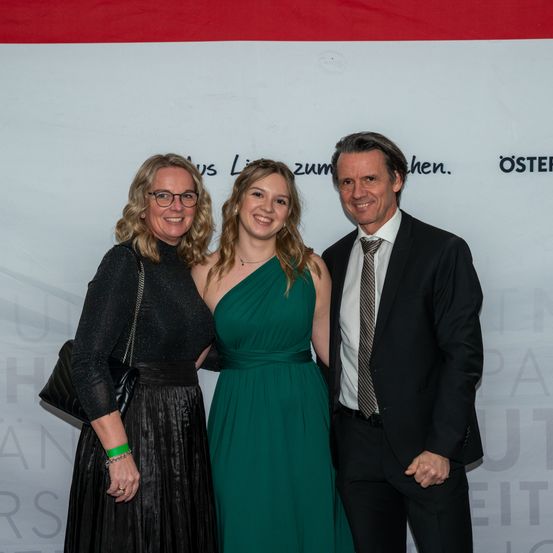 Drei Personen, zwei Frauen und ein Mann, stehen eng beieinander für ein Foto. Die Frauen tragen Kleider, eines grün und eines schwarz. Der Mann trägt einen Anzug und eine Krawatte. Sie lächeln in die Kamera.