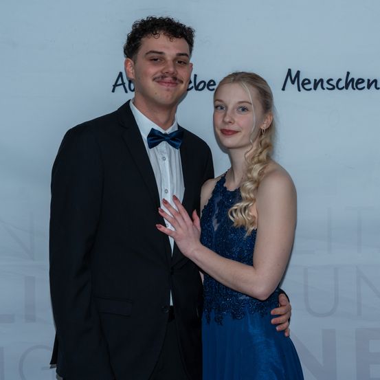 Ein Mann und eine Frau stehen zusammen und lächeln für die Kamera. Der Mann trägt einen schwarzen Anzug und eine blaue Fliege, während die Frau ein blaues Kleid trägt. Sie stehen vor einer weißen Wand mit deutschem Text.