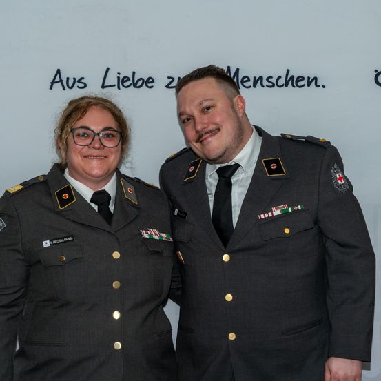 Zwei uniformierte Personen, ein Mann und eine Frau, stehen nebeneinander vor einer weißen Wand. Sie tragen beide eine Brille und haben Medaillen an ihren Uniformen. Hinter ihnen befindet sich eine Wand mit dem deutschen Text 'Aus Liebe zu Menschen.'