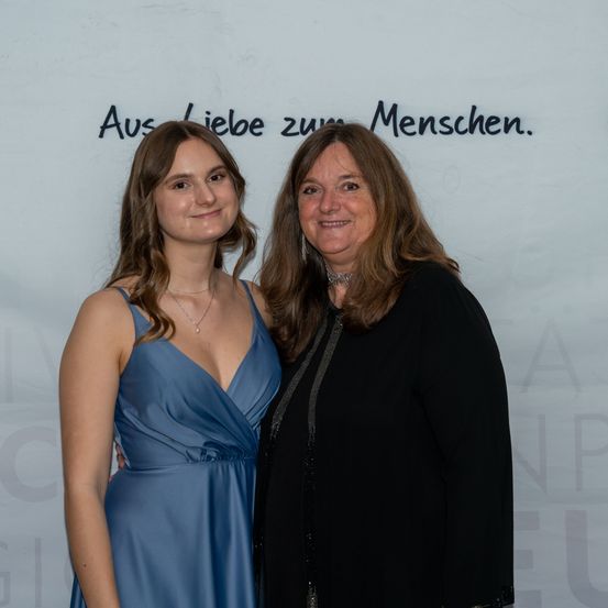 Zwei Frauen stehen nebeneinander und lächeln für ein Foto. Die Frau auf der linken Seite trägt ein blaues Kleid und die Frau auf der rechten Seite trägt ein schwarzes Oberteil. Hinter ihnen ist eine weiße Wand mit den Worten 'Aus Liebe zum Menschen'.